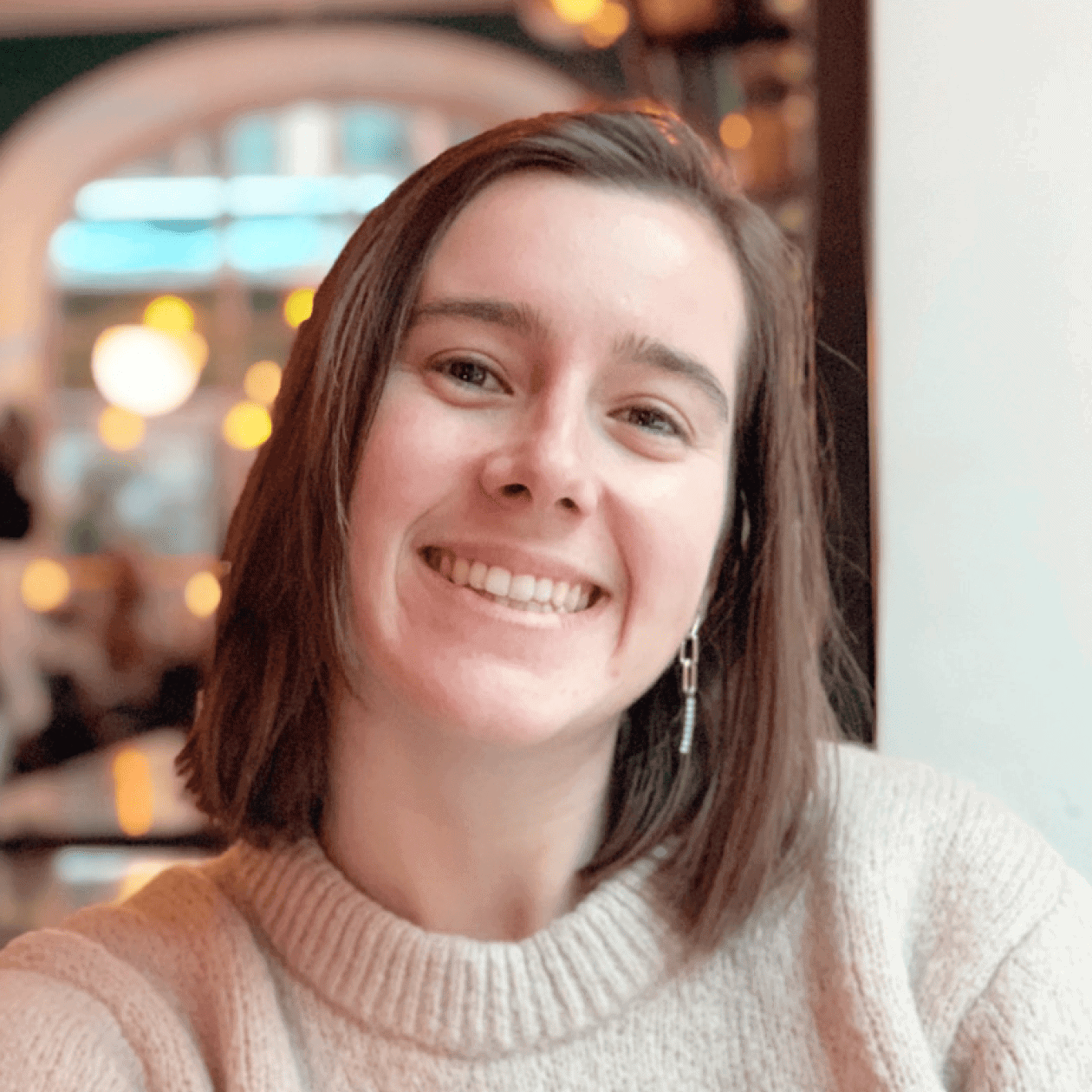 A cheerful individual with shoulder-length brown hair, dressed in a beige jumper and earrings, sitting indoors with cosy warm lighting in the background.