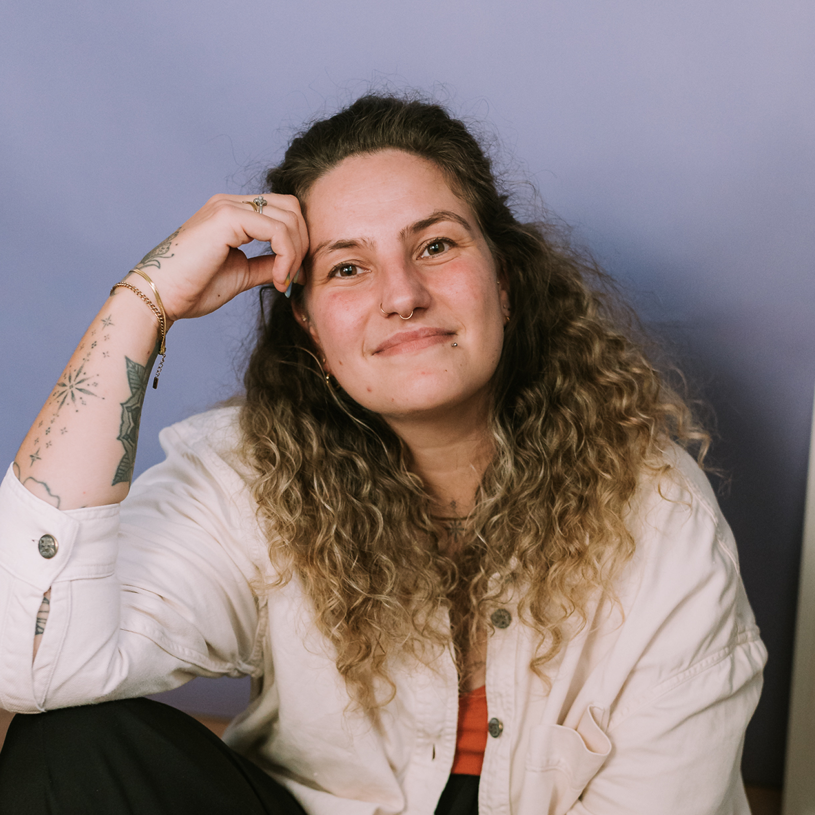 Trans non-binary designer with long curly hair and arm tattoos, wearing a light shirt over red top, smiling in front of purple background.