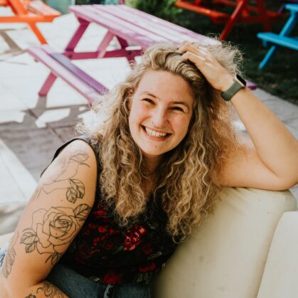 A cheerful person with curly hair and floral tattoos on their arm, seated on an outdoor bench. Brightly coloured picnic tables can be seen in the background. They are dressed in a floral top and wearing a watch.