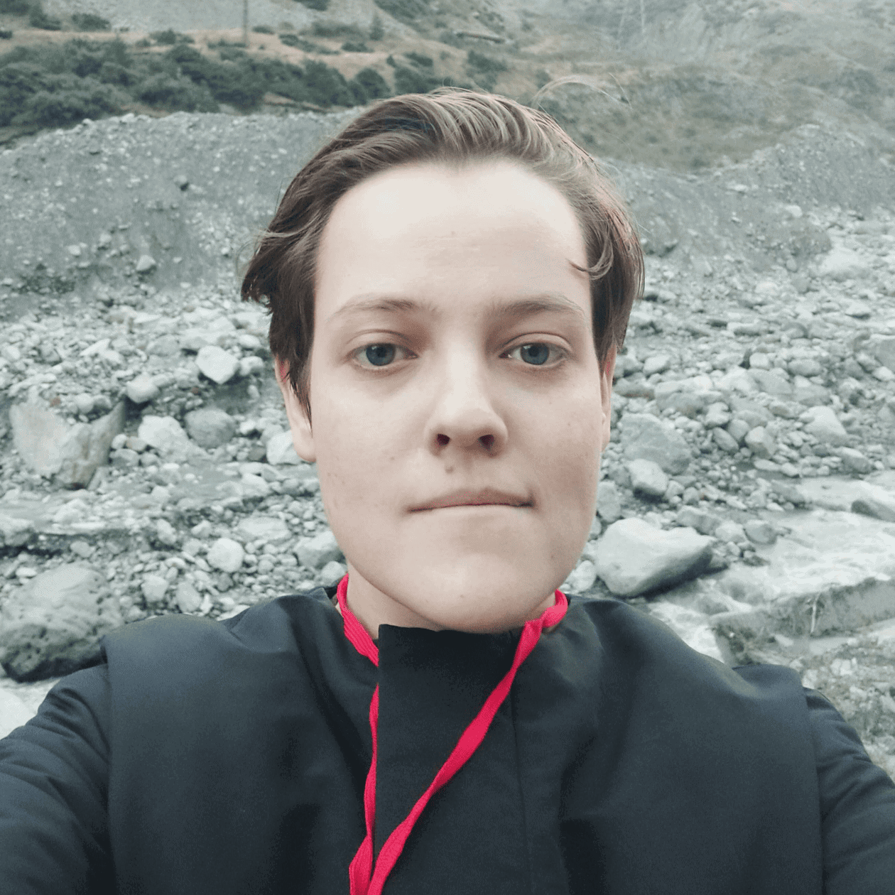 A person with short hair takes a selfie outside, dressed in a black top with a red tie. The backdrop features a rocky landscape with scattered vegetation.