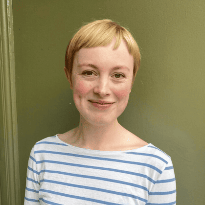 A person with short blonde hair smiling at the camera, dressed in a white and blue striped shirt. Background is plain olive green.