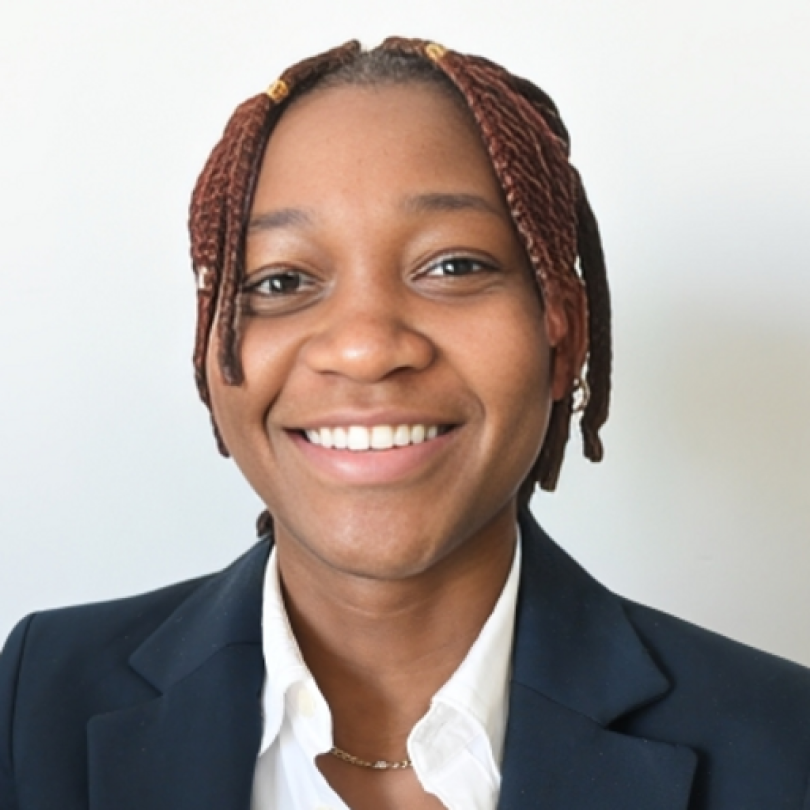 Cheerful individual with braided hair, donned in a dark blazer and white shirt, set against a simple backdrop.