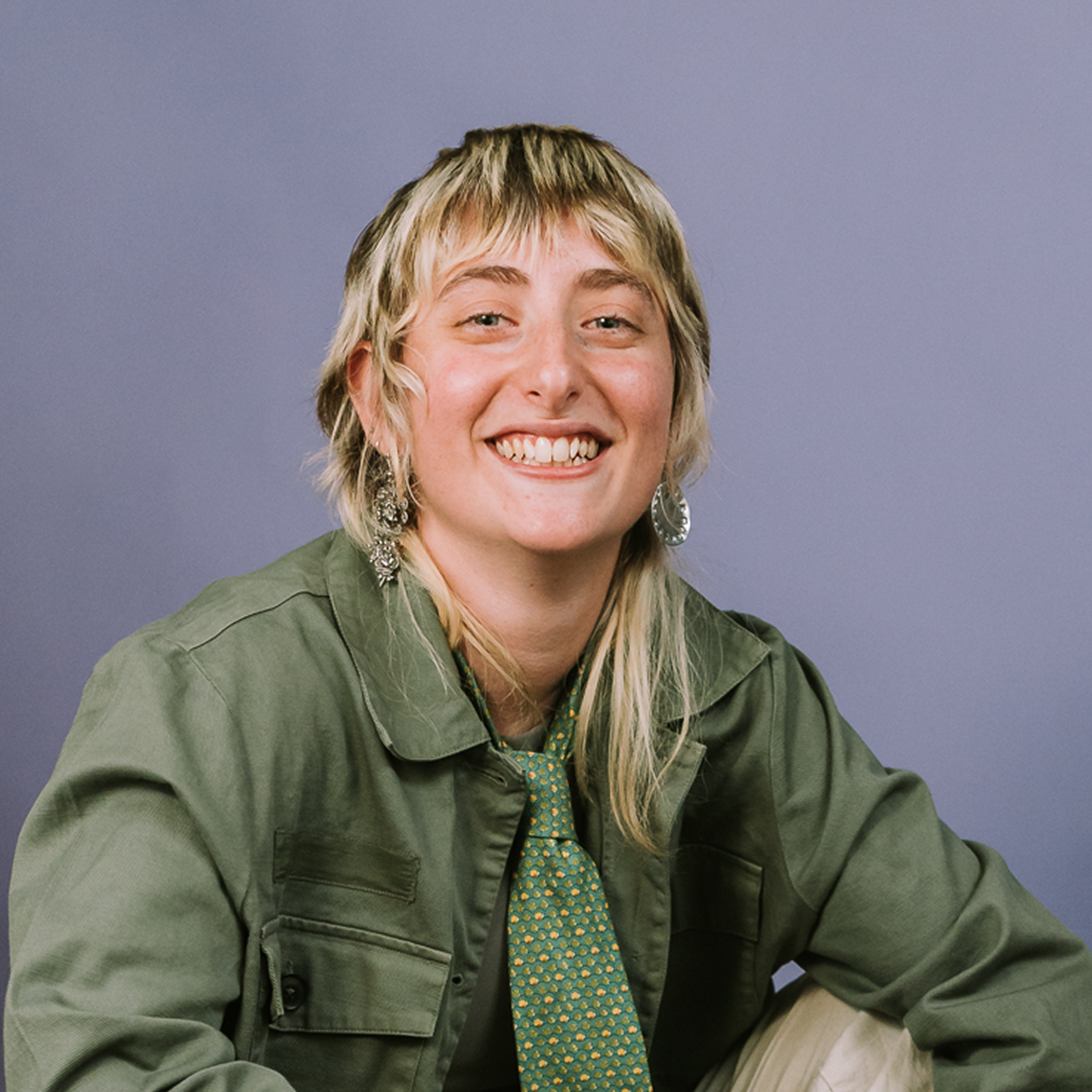 Queer nonbinary person with shaggy blonde hair, green jacket, patterned tie, and big earrings smiling against a plain purple background.