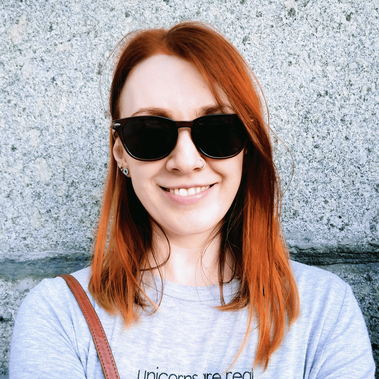 A smiling woman with vibrant red hair and sunglasses, wearing a grey shirt, stands in front of a textured grey stone backdrop.