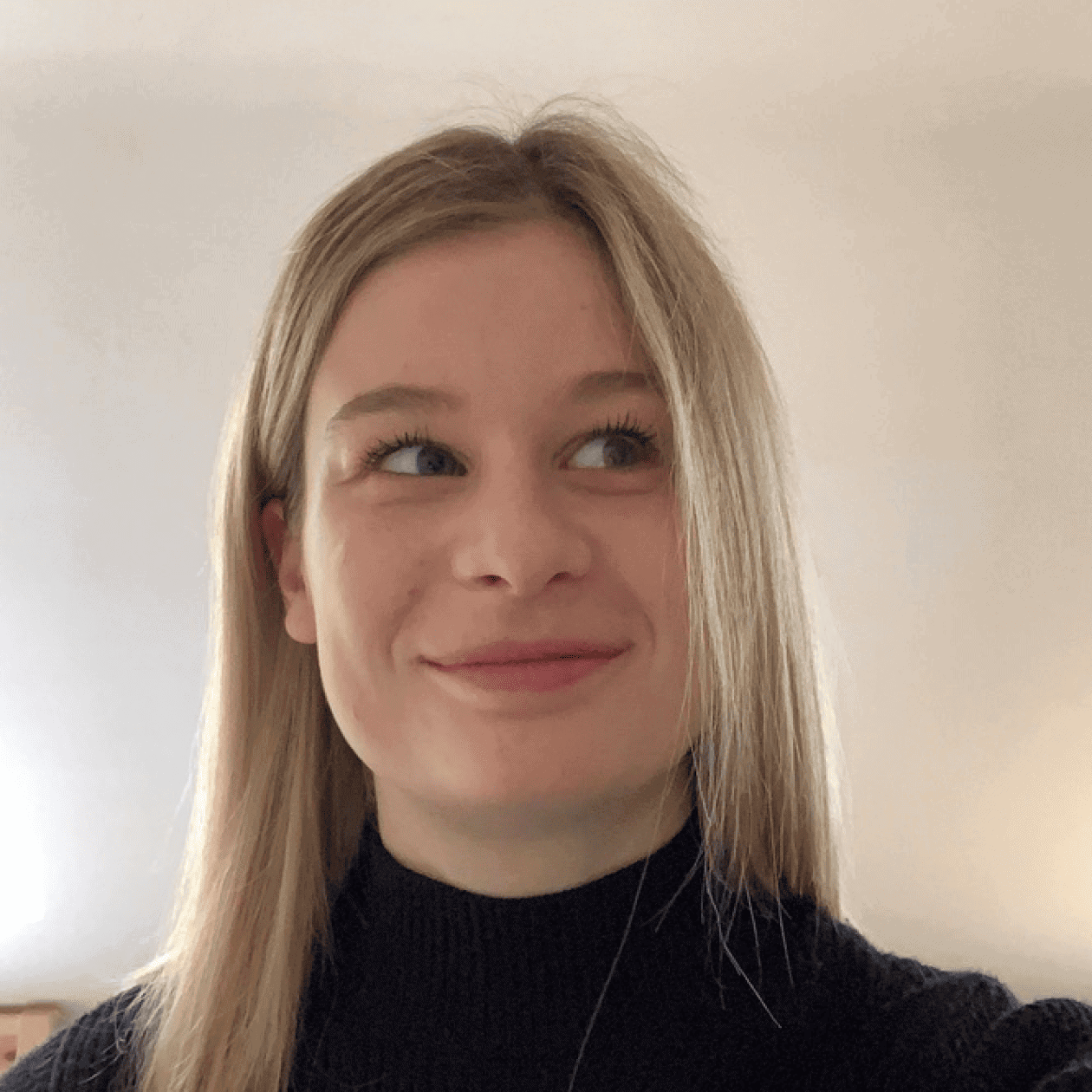 A person with long blonde hair in a black jumper smiles gently to the side against a simple, light-coloured background.