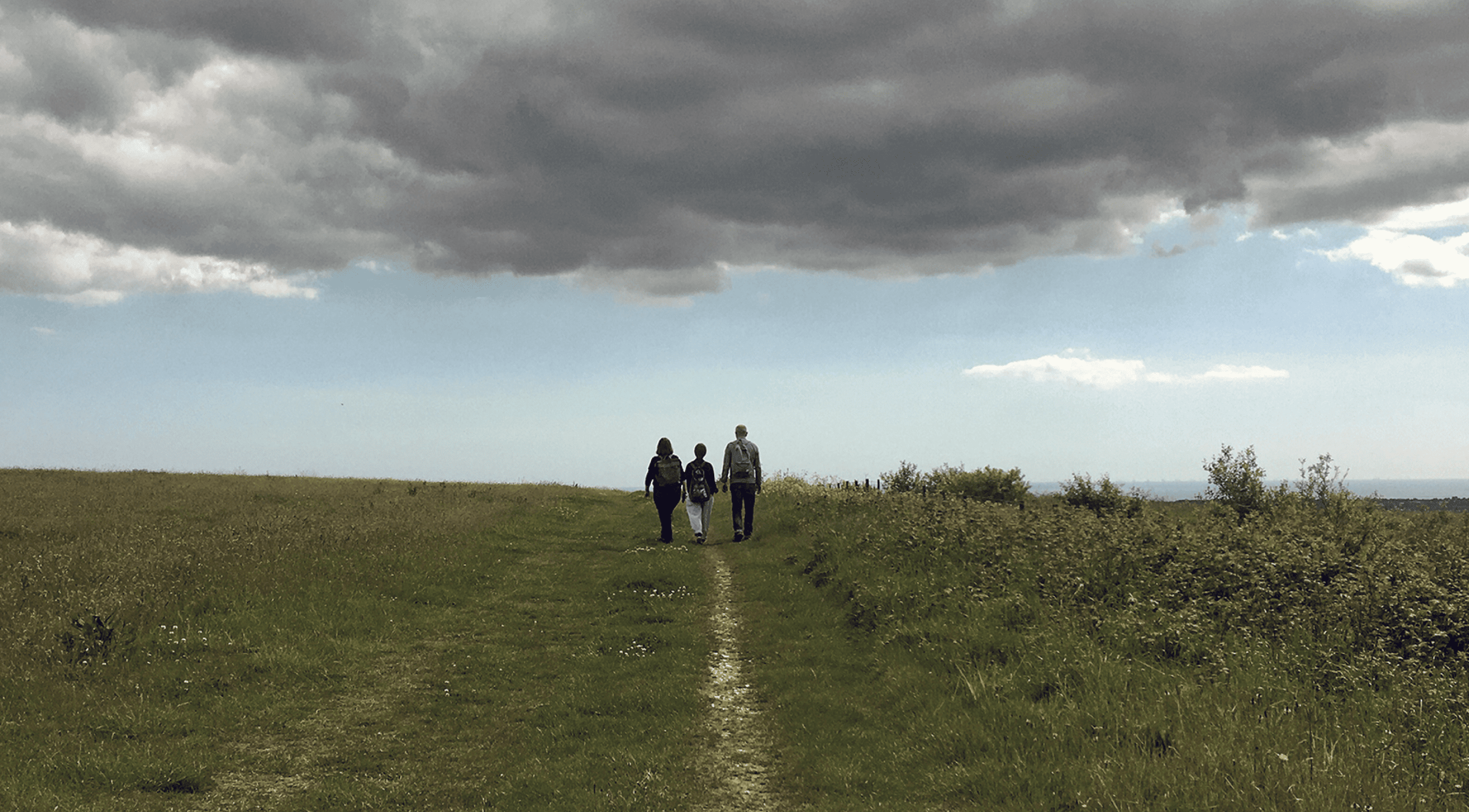 Three cheerful people strolling down a grassy path, flanked by expansive green fields. Overhead, the sky is a swirling mix of dark clouds and bright blue patches, creating an idyllic outdoor scene.