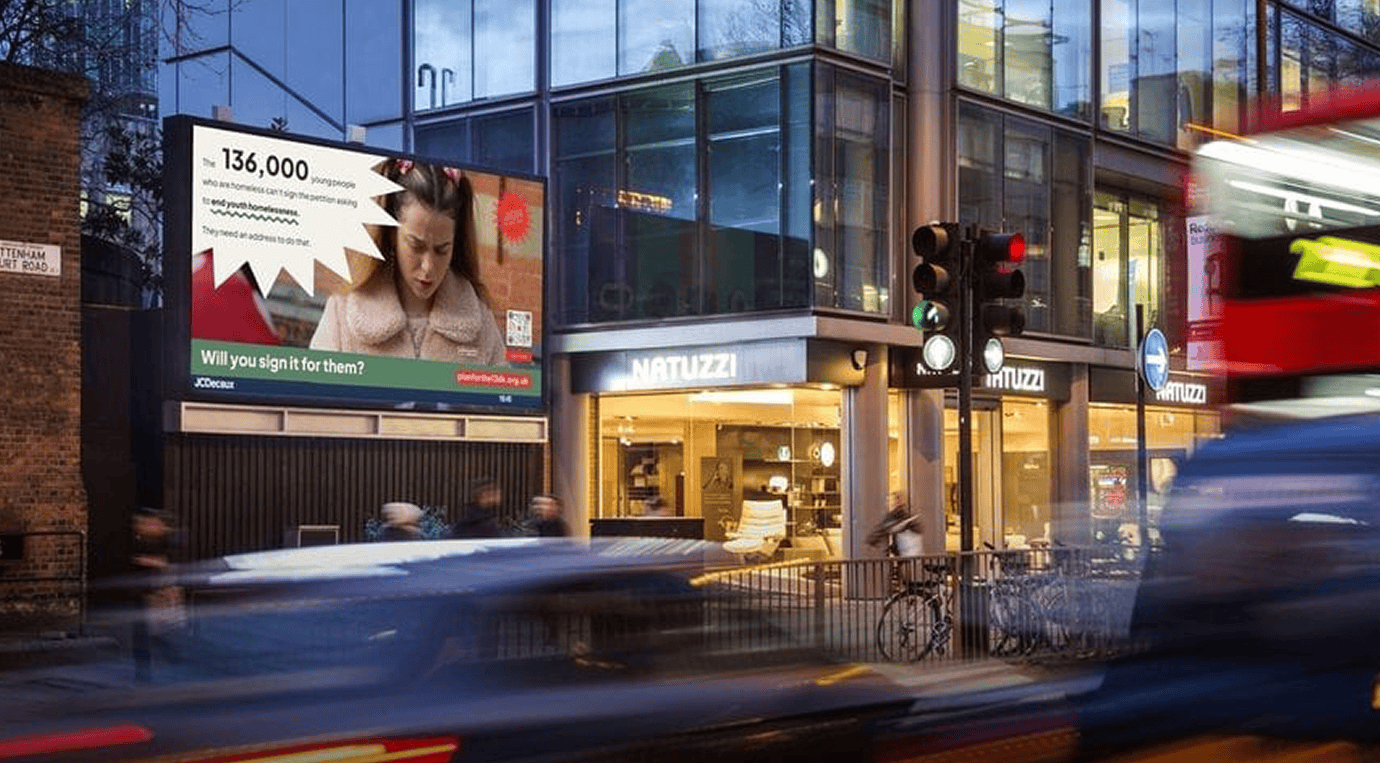 A bustling street scene with vehicles in motion blur. A digital billboard shows 136k no known pregnancies, advocating a signature campaign plan. The Natuzzi shop is prominently visible in the background.