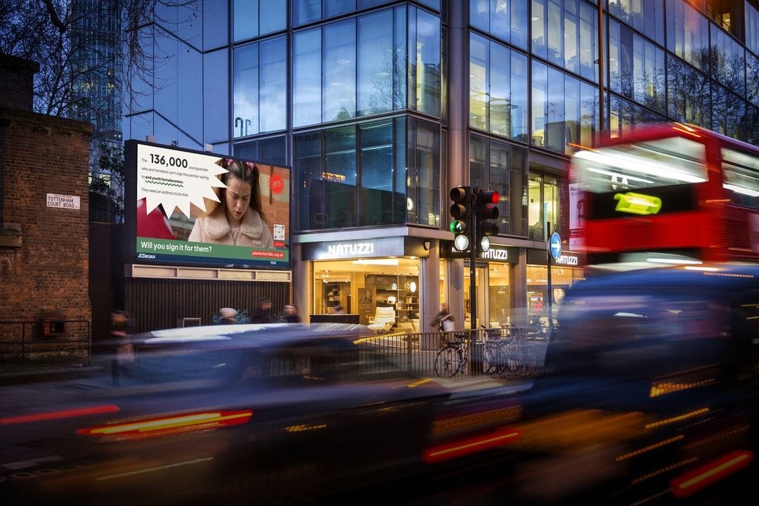 A bustling street with a blurred red double-decker bus zooming past. In the background, a modern glass building showcases a large digital billboard advertising a woman holding a mobile phone, with various shops behind the glass facade.
