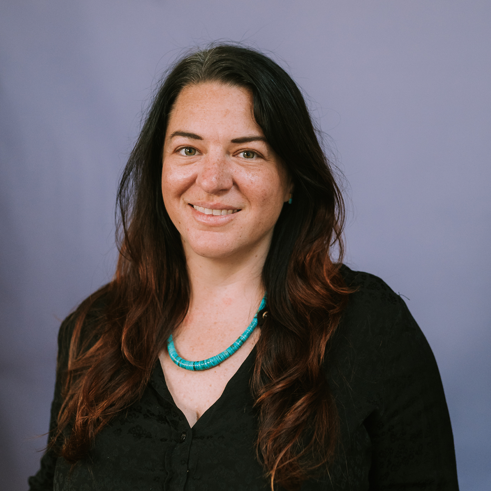 Ceysa McKechnie with long brown hair wears a black top and turquoise necklace, smiling in front of a plain purple background.