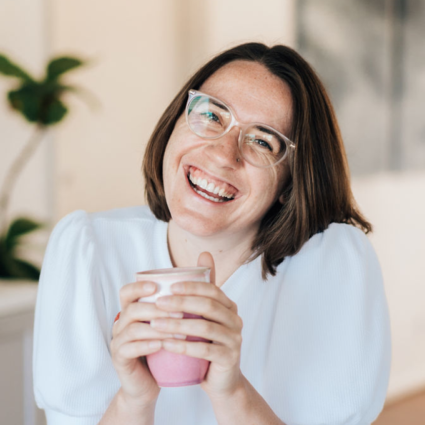 A cheerful person with shoulder-length brown hair and glasses holds a light pink cup, wearing a white blouse indoors. A blurred green plant is visible in the background.