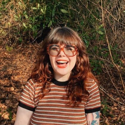 Rachel Wilson, a person with long, wavy red hair and large tortoiseshell glasses, smiles broadly. She is wearing a striped shirt while standing in a sunlit, wooded area. Dry leaves are scattered underfoot and lush green foliage fills the background.