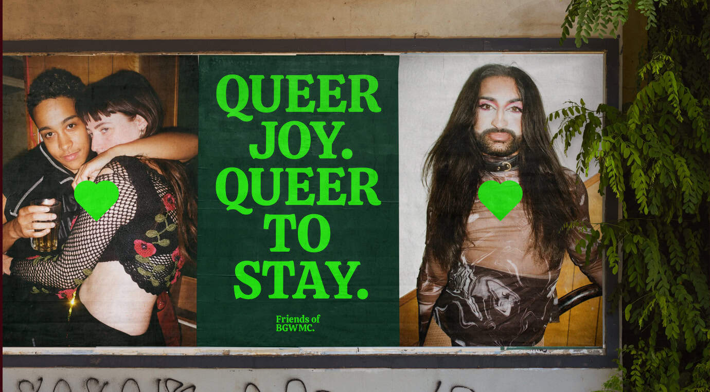 A large outdoor poster in Bethnal Green displays three individuals. On the left, two people embrace warmly; on the right is a person with long hair and a beard. Bold text declares, QUEER JOY. QUEER TO STAY. The poster includes green heart motifs, capturing the vibrant community spirit often found at Working Mens Club events.