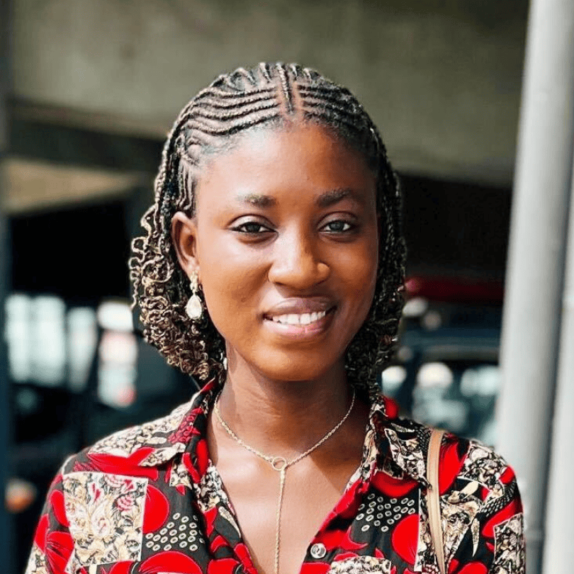 Amina Yakubu, with short braided hair, wearing a vibrant red and black patterned shirt, stands outdoors. Her radiant smile is highlighted against a blurred background.