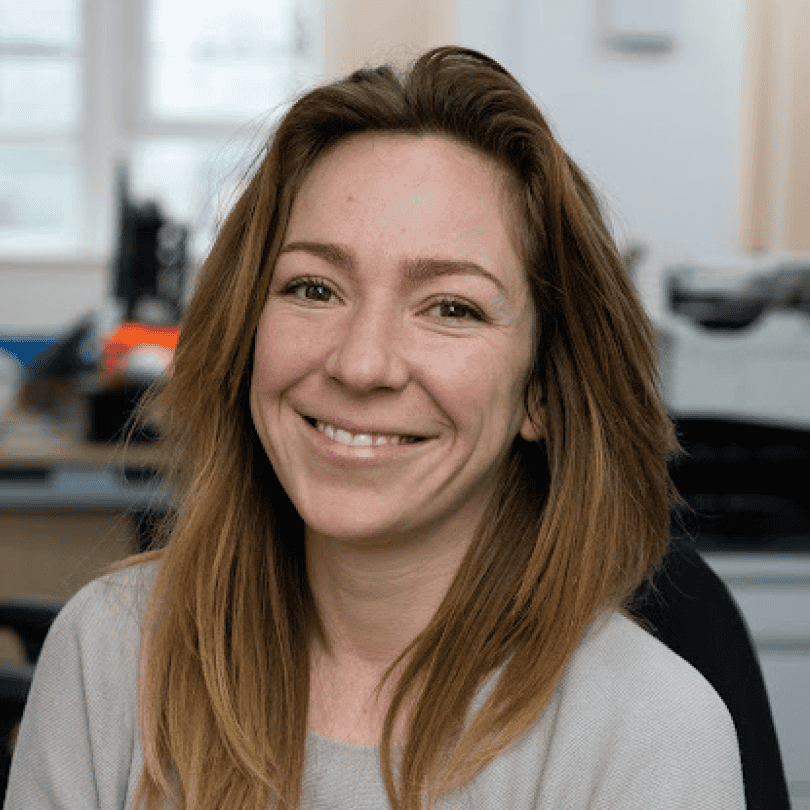 Meghan Mooney, smiling brightly with long brown hair, is seated in an office setting. She is wearing a light grey jumper against a softly blurred background.