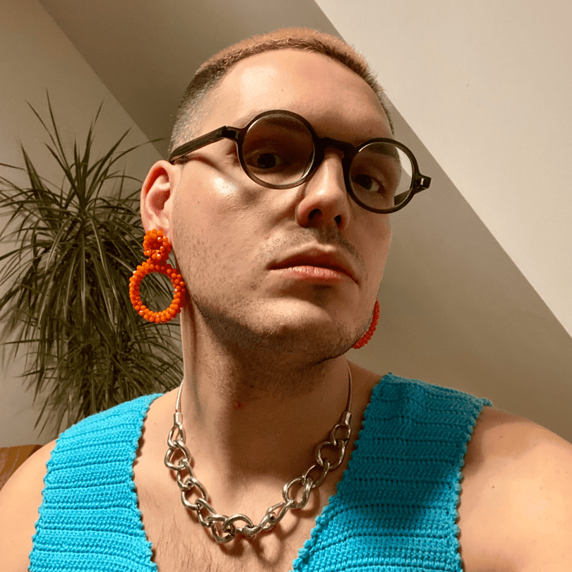 A person with round glasses, bright orange beaded earrings, and a chunky silver chain necklace is confidently facing the camera, wearing a knitted turquoise top. A tall plant is in the background.