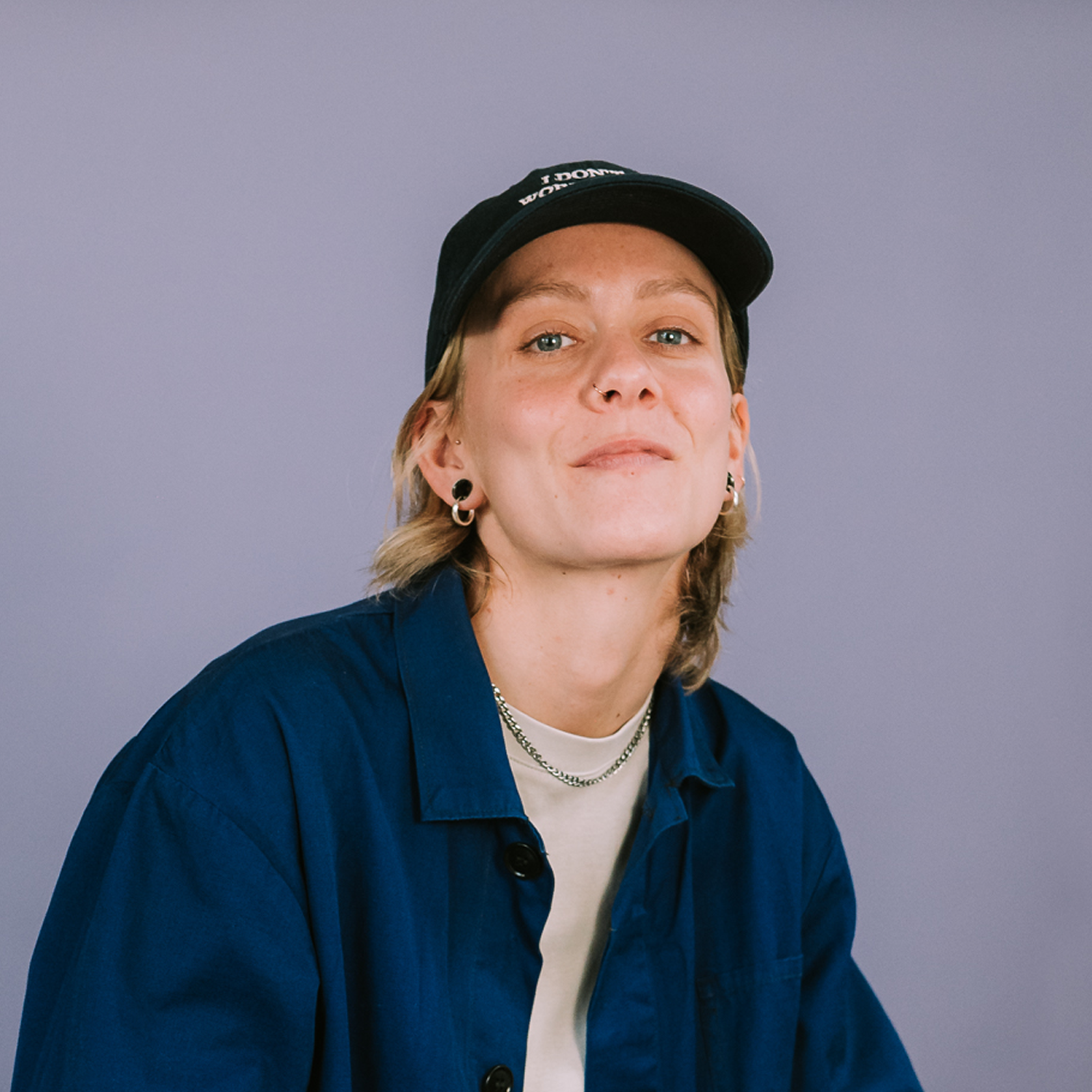 A person with short blond hair, a dark blue cap, a blue shirt, white tee and a silver chain necklace smiles at the camera, on a light purple background.