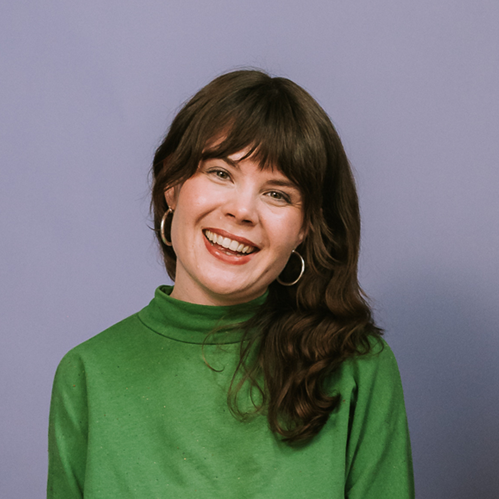Smiling person with long brown hair, green turtleneck and hoop earrings, stands confidently before a plain purple background.