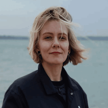 Danielle Mustarde, a woman with short blonde hair wearing a black jacket, stands outdoors beside a body of water on a windy, cloudy day.