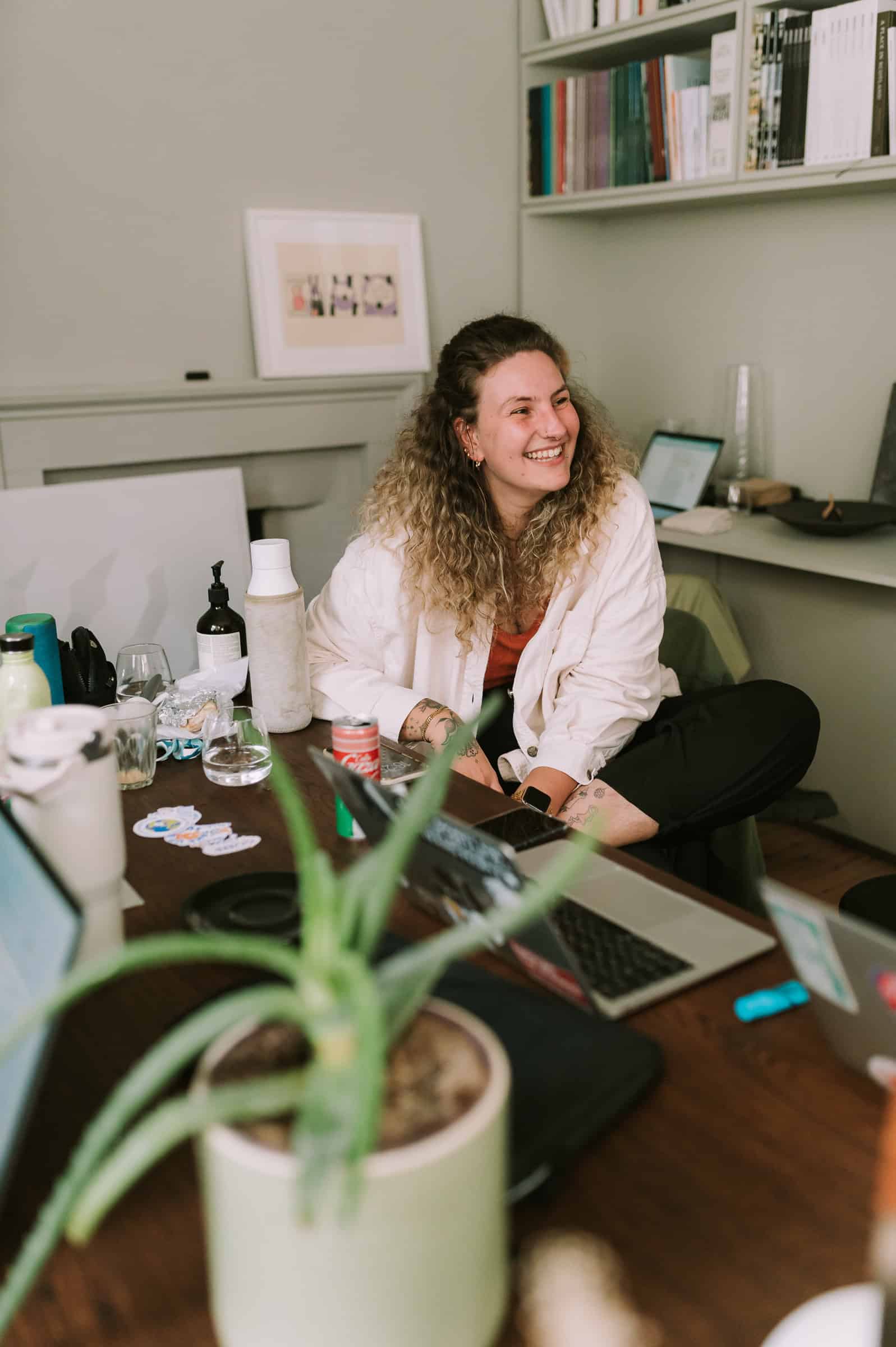 Cecilia Righini, curly-haired and wearing a light shirt, smiles at a desk with laptops, plant, water bottles, and bookshelves behind.
