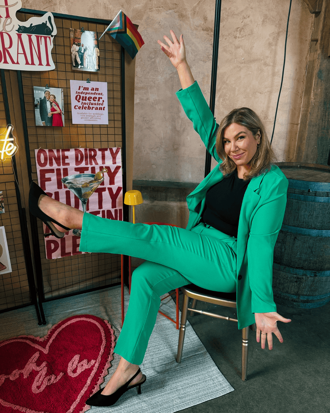 Smiling woman in bright green suit sits on chair with leg raised, arms up; pride flags, signs, barrel and heart-shaped rug around her.