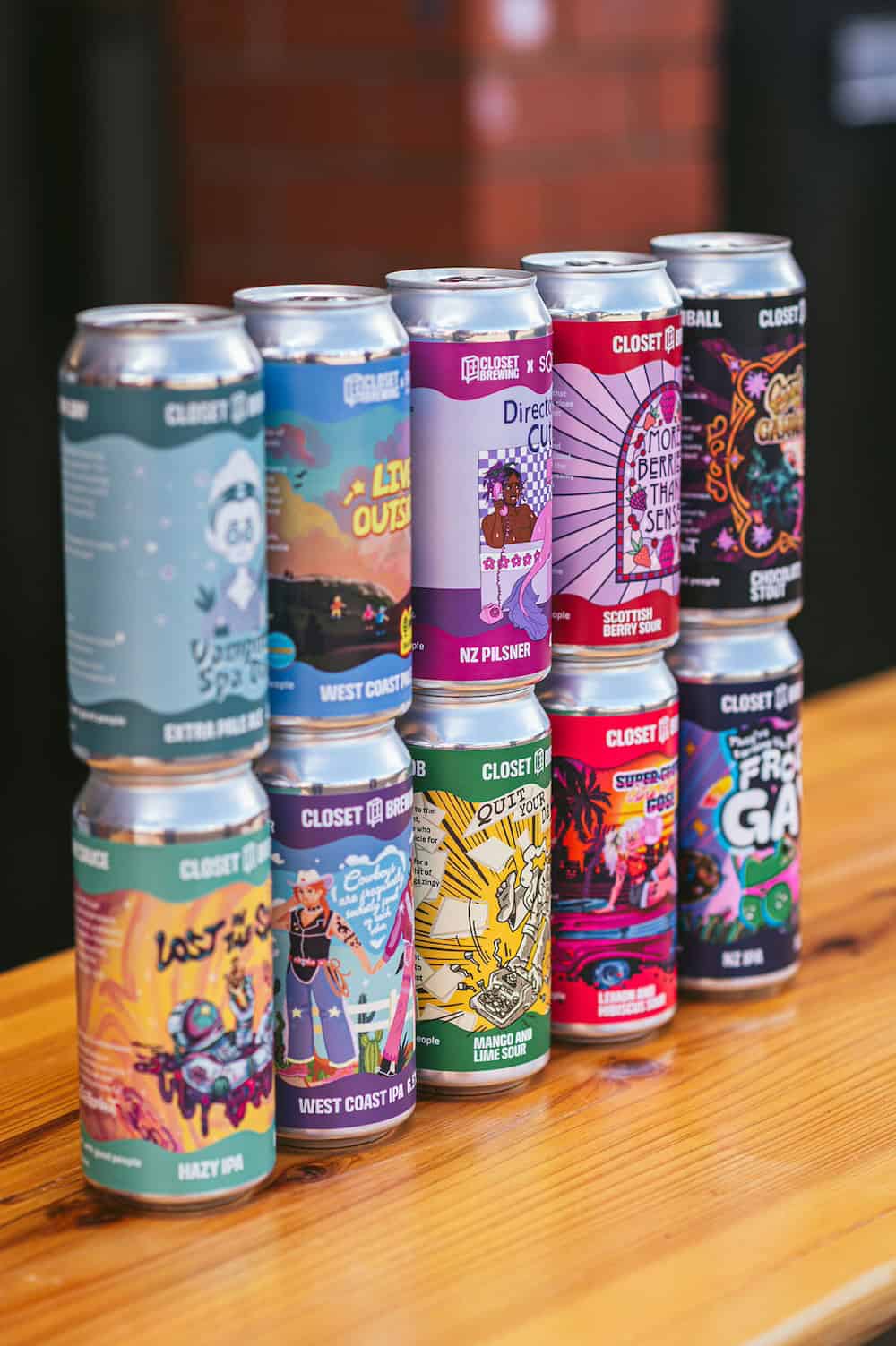 Nine colourful Closet Brewing craft beer cans with bold labels stacked in three rows on a wooden table, blurred indoor background.