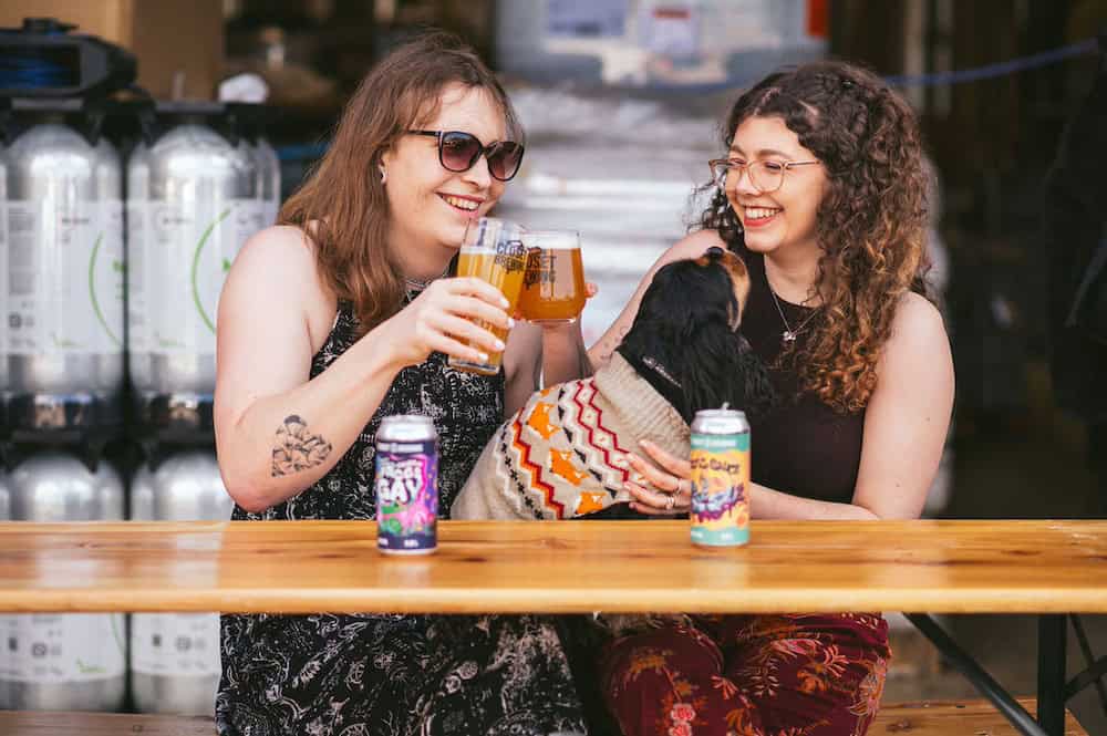 Two women enjoy Closet Brewing beers outdoors at a wooden table, smiling, with a small dog in a jumper and two beer cans between them.
