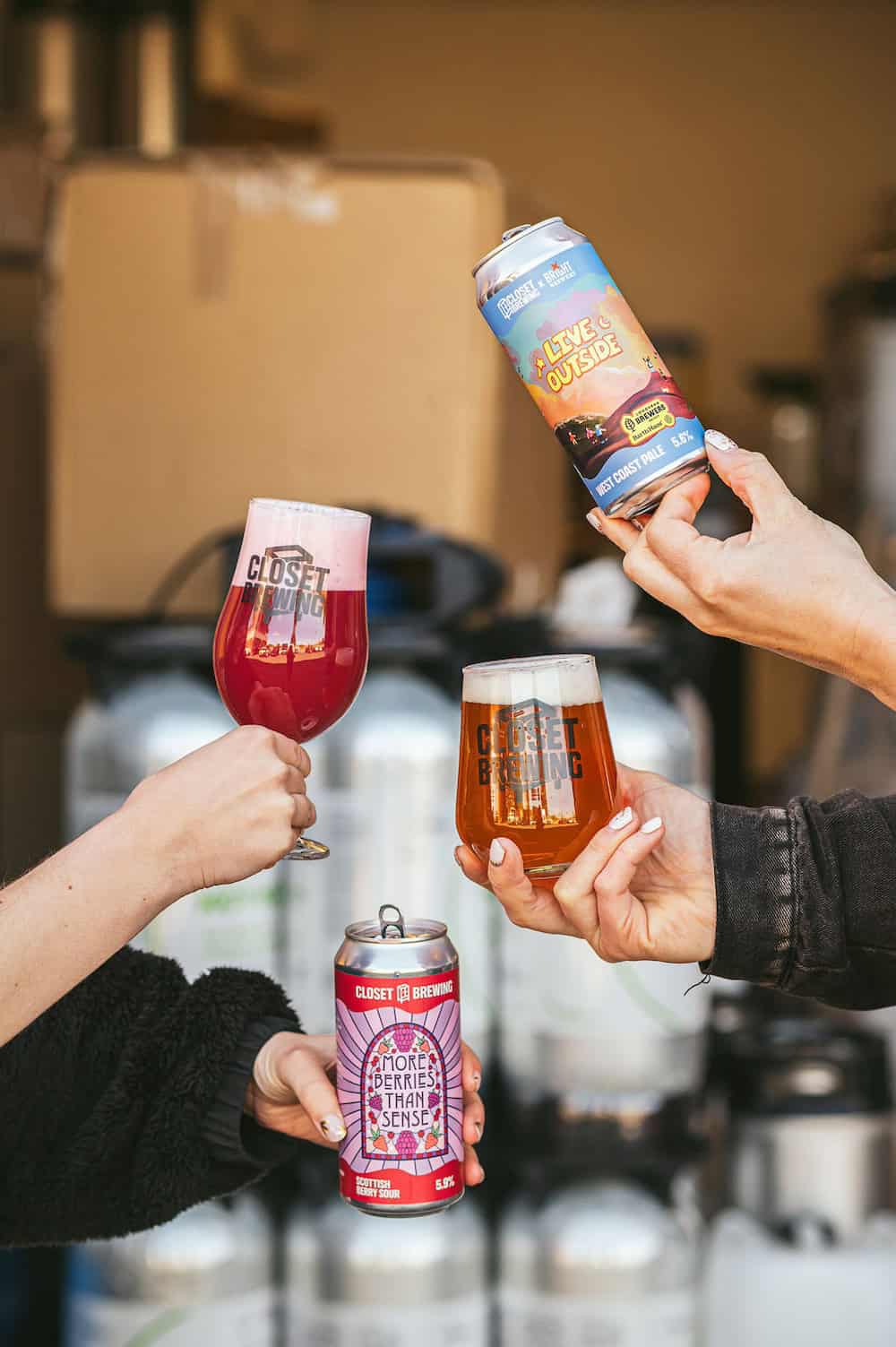 Three hands raise Closet Brewing beer cans and glasses—one pink, one amber—against stacks of boxes and metal kegs in a brewery.