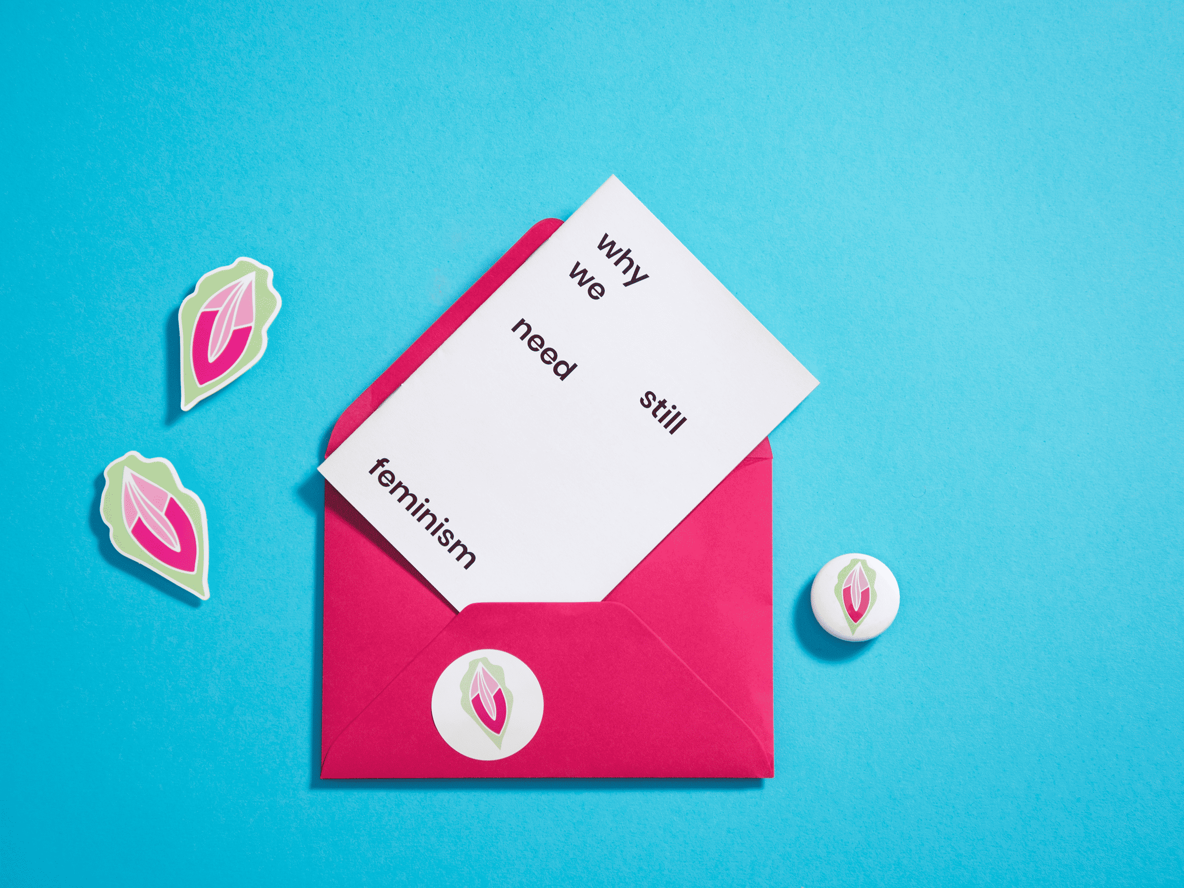 Pink envelope with vulva sticker holds a note saying 'why we still need feminism', plus two vulva stickers and matching badge on blue background.