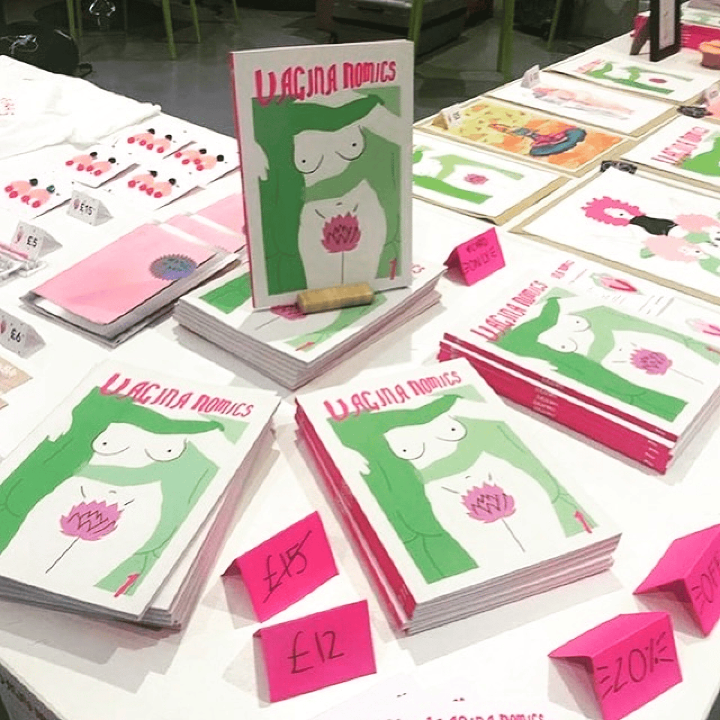 A table with piles of Vagina Nomics comics showing a female torso and pink flower, pink price tags, plus colourful zines and art.