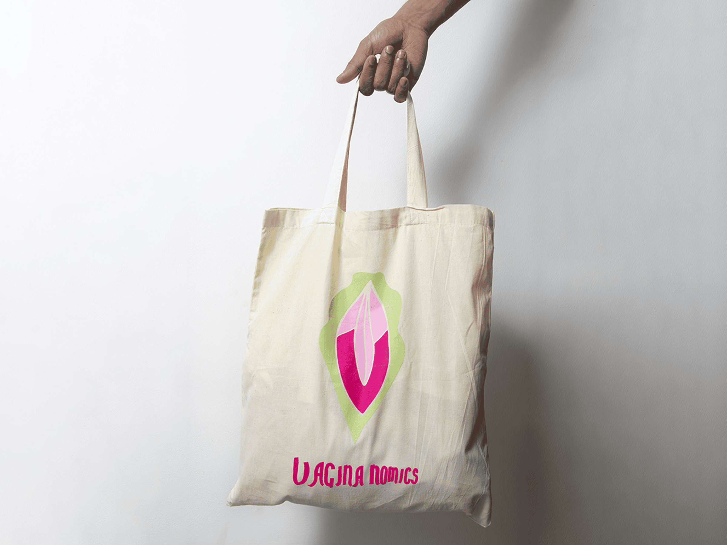 Hand holding a beige tote bag with pink and green vulva design and "Vagina Nomics" text, against a plain white background.