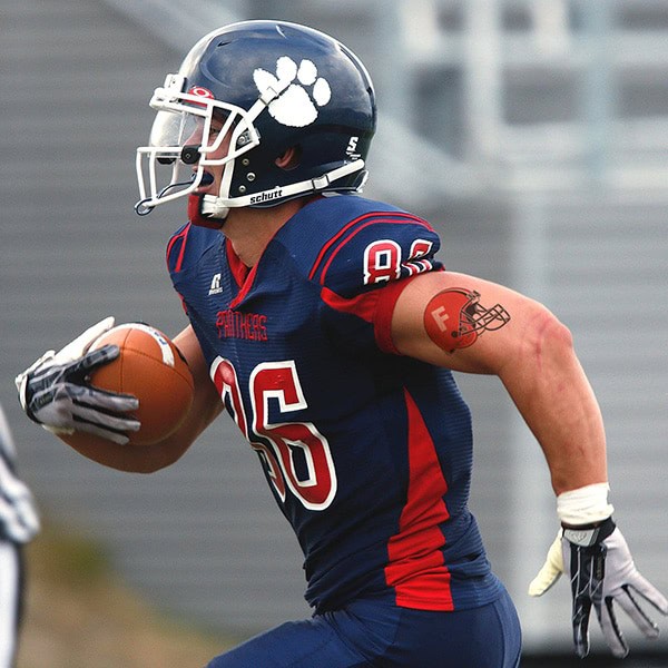 Atleta de fútbol americano con casco y uniforme, mostrando un estampado de impresión en vinilo, en acción durante un partido. Equipo con logotipo y detalles en letras, ejemplo de uso de vinilos para personalización deportiva.