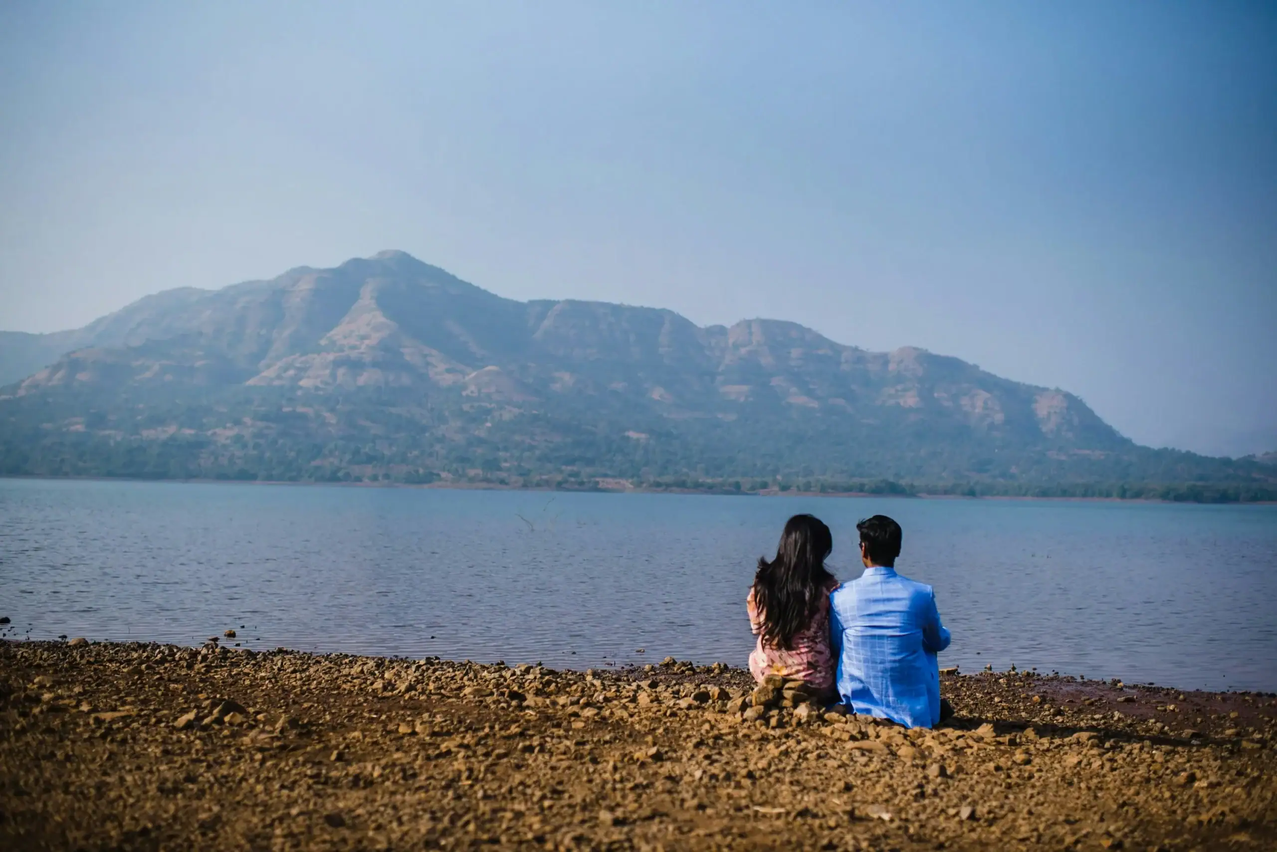 Couple sitting by a tranquil lake, gazing at distant mountains under a clear blue sky.