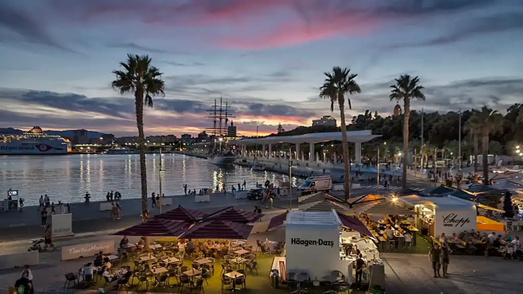 Ambiente nocturno en Muelle Uno, Málaga: farolas, reflejos sobre el agua y gente paseando.