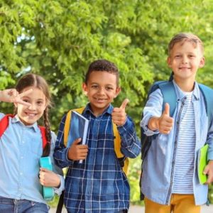 Friendly primary school children walking outdoors, smiling with backpacks, in a green schoolyard.