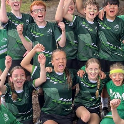 School children celebrating outdoors at Mangorei School in New Zealand, wearing green sports jerseys, smiling, raising fists in victory, team spirit, school sports event, kids in sports gear, happy students, community, youth activity, NZ education, outdoor sports, group achievement, school pride, team unity, youth sports.
