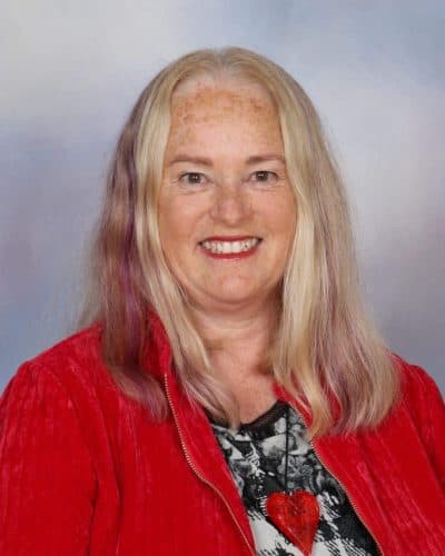 Bright smiling woman with long blonde hair wearing a red jacket, standing in front of a plain background.