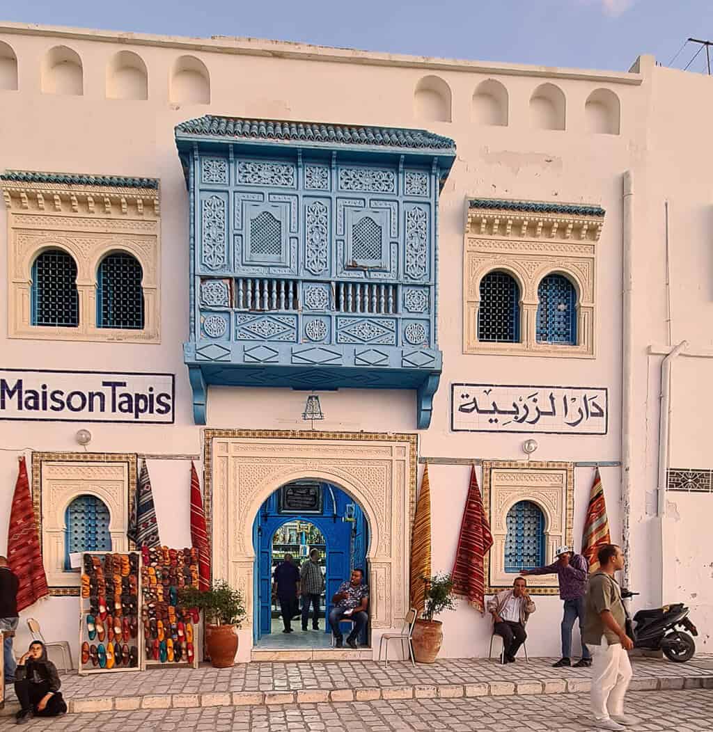Traditional carpet shop facade in Kairouan, Tunisia, showcasing ornate Arabic architecture with intricate blue wooden balcony and arched windows. The entrance displays colorful handmade rugs and slippers, reflecting Tunisia’s rich artisanal culture and the city’s reputation as a hub for traditional crafts near the Great Mosque of Kairouan.
