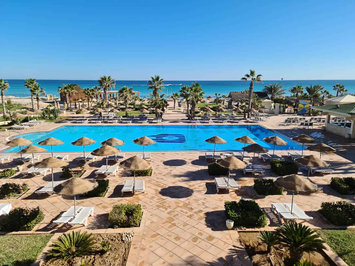 Luxurious beach resort on Djerba Island, Tunisia, featuring a large outdoor pool surrounded by sunbeds, thatched umbrellas, and lush palm trees with a view of the turquoise Mediterranean Sea. This picturesque vacation destination is ideal for travelers seeking relaxation, beachfront accommodation, and all-inclusive amenities on the island of Djerba.