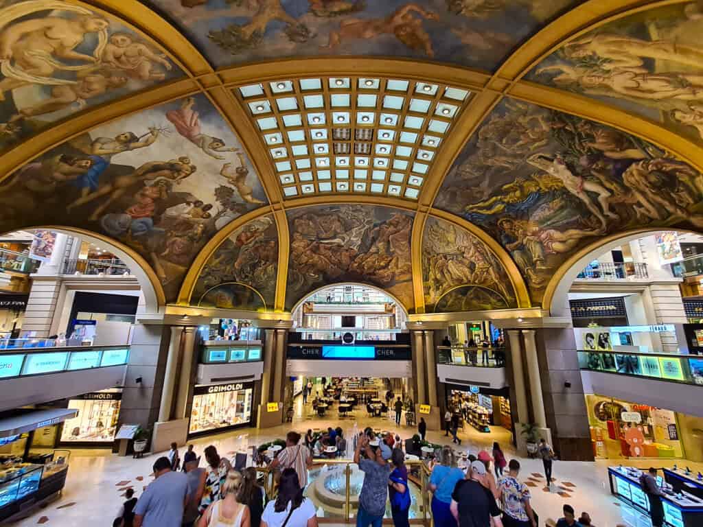 This image showcases the magnificent interior of the Galerías Pacífico shopping center in Buenos Aires. The ceiling is adorned with elaborate frescoes and golden arches that give the space a luxurious feel, reminiscent of a grand art gallery. The natural light streaming in from the skylights above illuminates the artwork and the bustling shoppers below, highlighting the building's fusion of commerce and art.