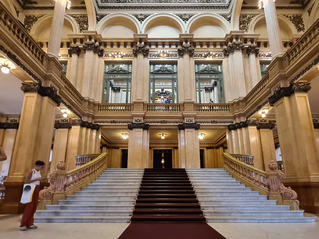 The grand staircase inside the Teatro Colon Buenos Aires. The opulent interior features marble steps, flanked by ornate balustrades and classical Corinthian columns. The ceiling boasts elegant stained glass and detailed plasterwork.