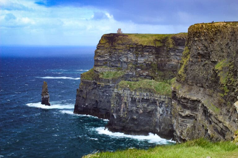 The photo showcases the majestic Cliffs of Moher on the west coast of Ireland, with the Atlantic Ocean churning below. A solitary sea stack stands near the cliff base, separated from the mainland by centuries of erosion. The cliffs rise steeply from the ocean, their stratified layers of rock visible, topped with a lush green surface. Atop the cliffs, a small stone tower or building offers a lookout point for visitors, dwarfed by the scale of the natural landscape. The sky above is a mix of blue with wispy white clouds, indicating variable weather conditions. The powerful waves hitting the cliff base create white foam, highlighting the wild and natural beauty of this Irish landmark.