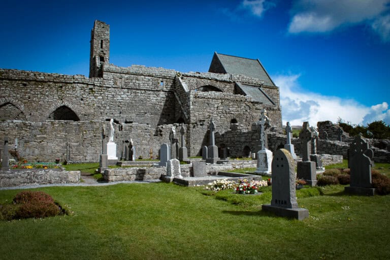 The photo features Corcomroe Abbey, an ancient Cistercian monastery ruin located in County Clare, Ireland. The remains of the abbey are built of grey stone, with sections of the walls still standing, showcasing the traditional Gothic arches and architectural details. Within the ruins, there's a well-kept graveyard with various gravestones, including traditional Celtic crosses and more modern headstones, one prominently labeled "RYAN." The grass around the graves is lush and green, indicating regular care. A statue of a religious figure stands within the grounds, adding a serene and sacred atmosphere to the site. The sky above is mostly clear with a few clouds, suggesting a bright and peaceful day.