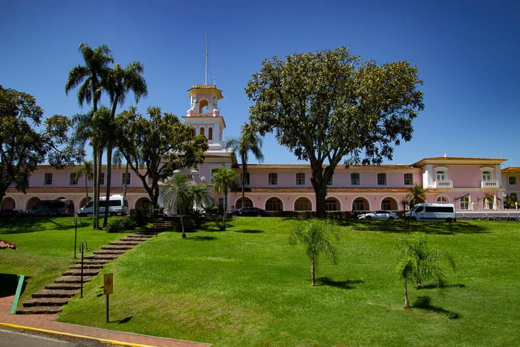 The photo showcases a picturesque two-story building painted in a soft pink hue with white trim, featuring a prominent clock tower with a spire at the center. The architecture suggests a colonial style with arched porticoes lining the front of the building. Lush, well-manicured lawns surround the structure, dotted with tropical palm trees and a large, shady tree at the forefront. The clear blue sky suggests a bright, sunny day. Stone steps lead up to the entrance, and a few vehicles are parked on the side, indicating that this may be a place of interest, possibly a hotel or historic site, which is frequented by visitors.