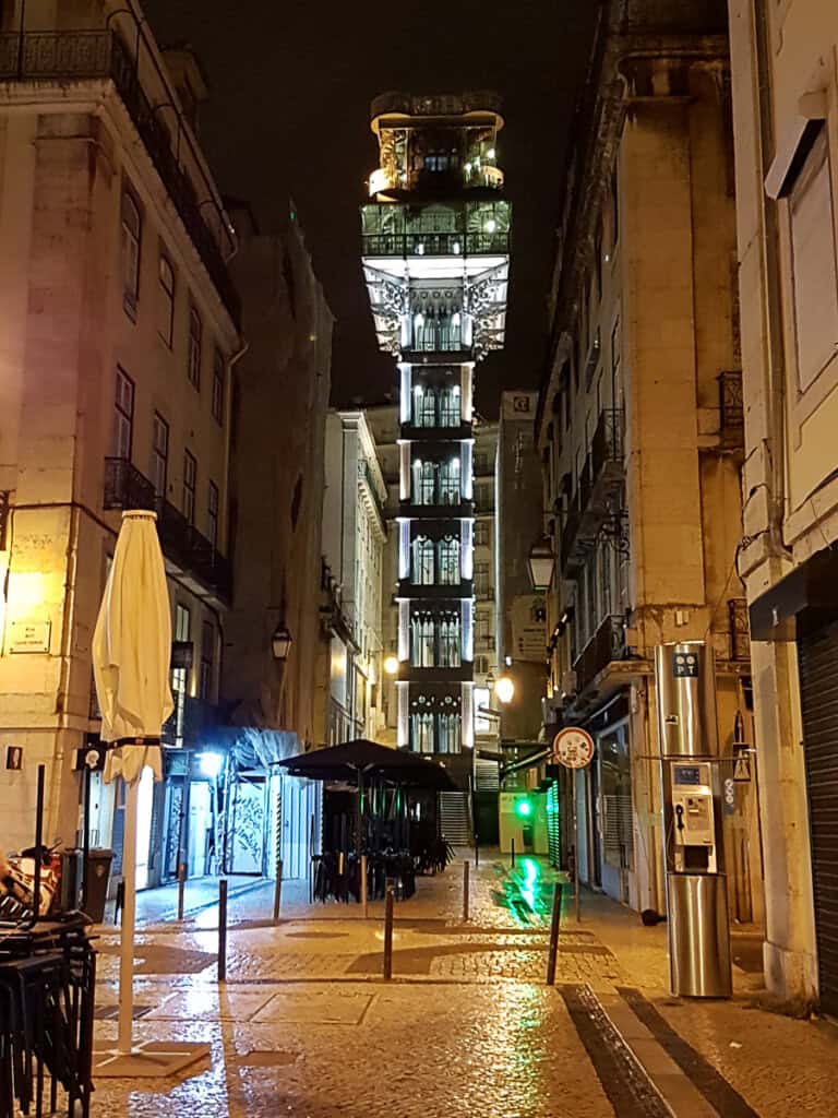 A night scene of the Santa Justa Lift in Lisbon, illuminated against the dark sky. The ornate, vertical elevator connects two streets and stands out amidst the closed shops in the quiet evening.
