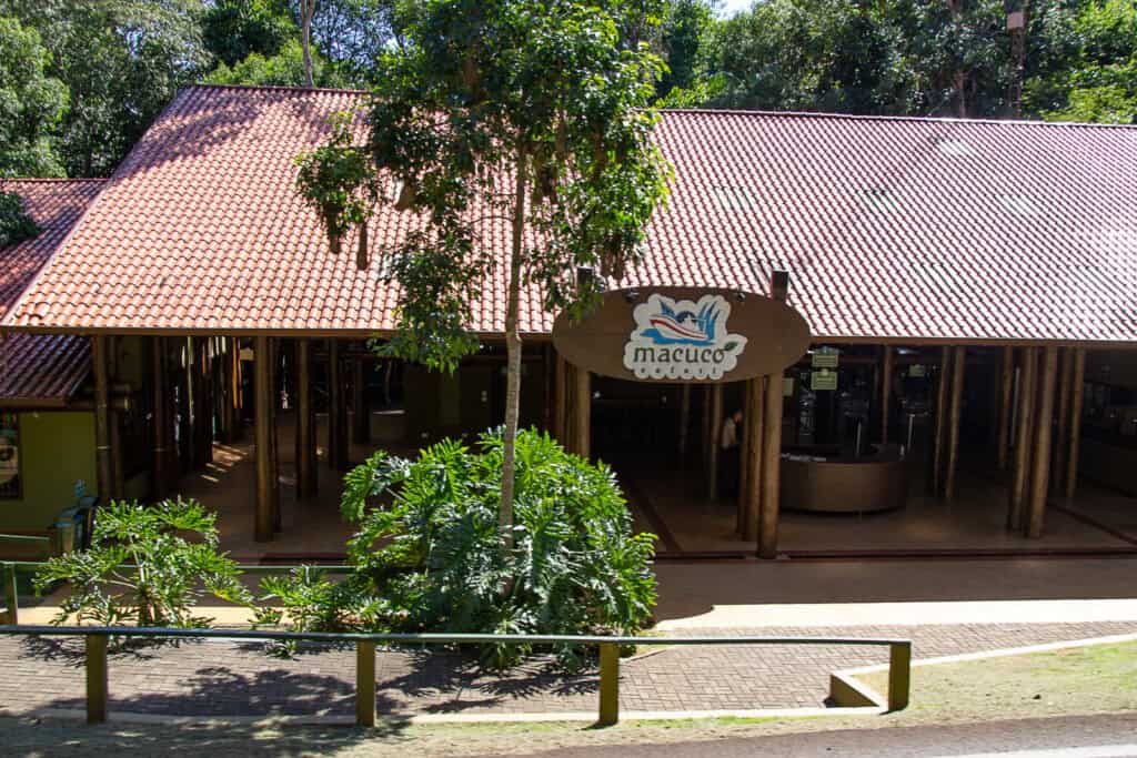 The photo shows a single-story building with a terracotta-tiled roof nestled among verdant greenery. A sign with the word "Macuco" and an illustration of a bird and leaves hangs prominently over the entrance, suggesting the building is a stop or attraction, possibly named Macuco, within a larger park or tour area. The structure has an open, pavilion-like design with large supporting wooden pillars, offering a welcoming area for visitors. The setting appears tranquil and shaded, a pleasant stop for those exploring the surrounding natural attractions. A clear sky and bright sunlight indicate a fine weather day, perfect for outdoor activities.