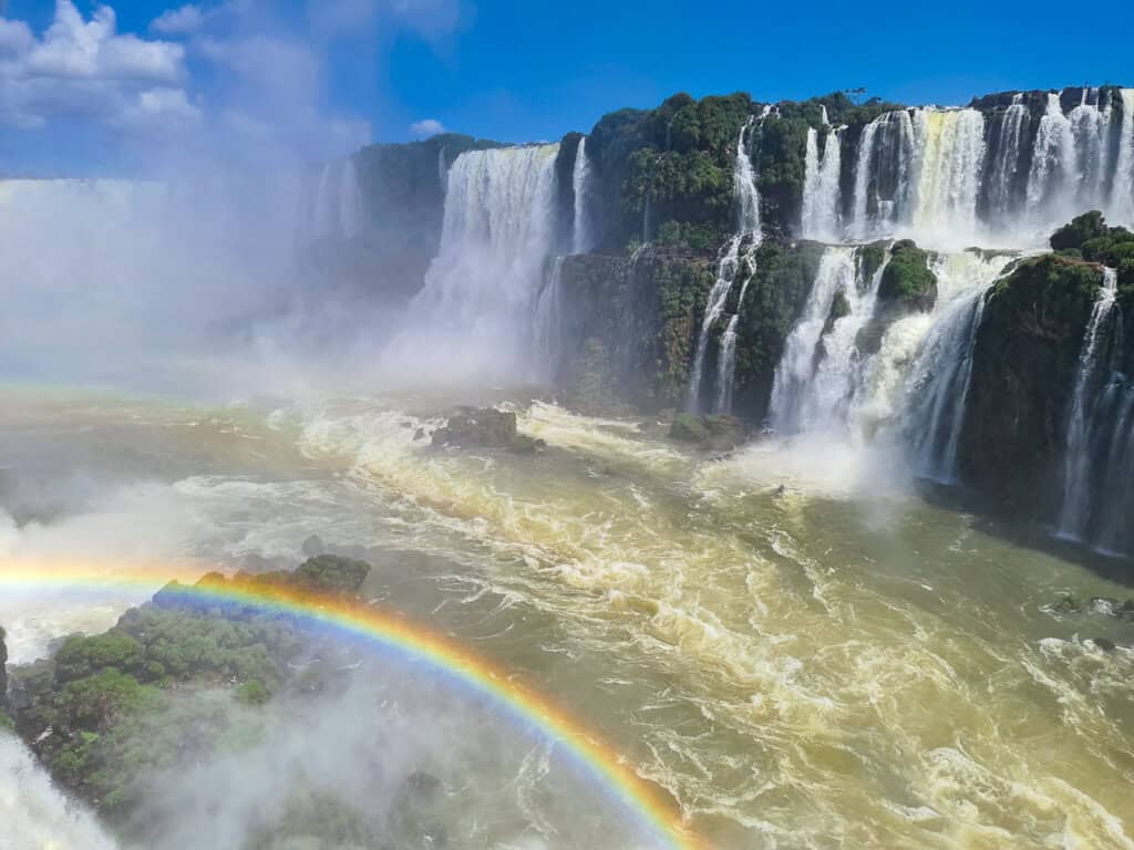 The photograph showcases a striking view of Iguazú Falls, where numerous waterfalls cascade into a turbulent, golden-brown river. A vivid rainbow arches gracefully through the mist created by the falls, adding a spectrum of color to the scene. The lush greenery of the surrounding forest is partially visible, framing the natural wonder, while the powerful rush of water conveys a sense of the falls' majesty and the dynamic energy of nature.