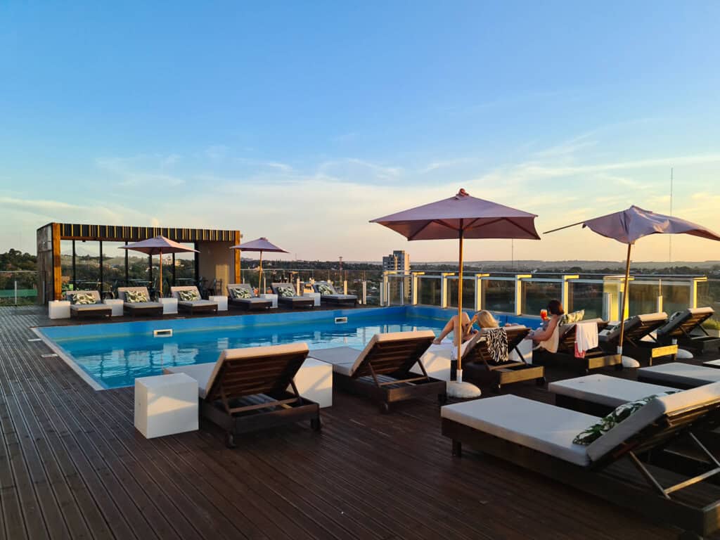 The image shows a luxurious rooftop swimming pool at dusk, with a clear blue sky above. The pool area is adorned with stylish sun loungers, some of which are occupied by guests enjoying the tranquil setting. Large umbrellas provide shade, and a chic bar structure offers refreshments. The wooden deck and glass balustrades add to the modern aesthetic, while the background reveals a sprawling landscape that fades into the horizon, highlighting the urban setting of the O2 Iguazu Hotel.