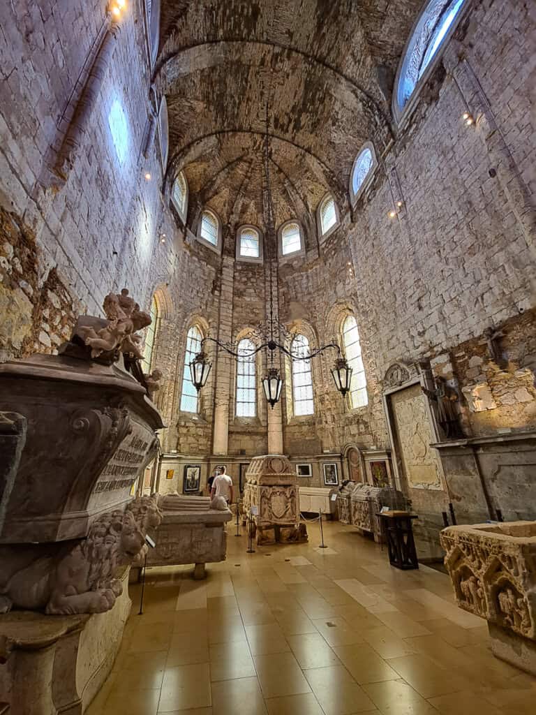 Inside the Carmo Archaeological Museum in Lisbon, the photo displays a collection of ornate sarcophagi set against the rugged stone walls of the Carmo Church, with a visitor in contemplation.
