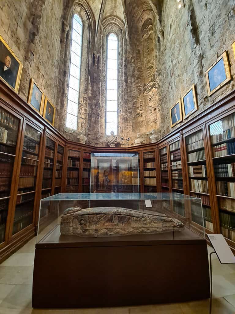 A wide-angle photo of the interior of the Carmo Archaeological Museum in Lisbon, with shelves lined with old books encircling a central glass case displaying an artifact, evoking a sense of historical depth.