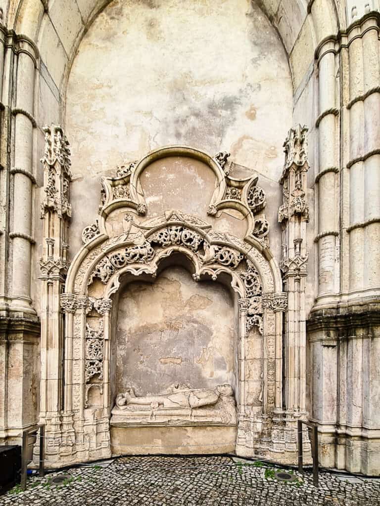 The photo showcases an ornate, intricately carved stone archway within the Carmo Convent in Lisbon. The weathered texture of the stone and the elaborate gothic tracery stand out against the faded wall. The cobblestone ground adds to the historical ambiance of the setting.